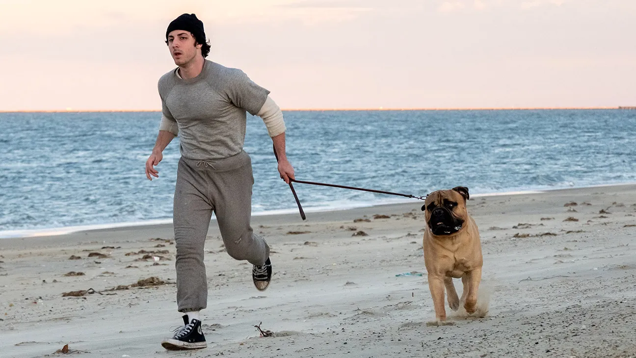Gri eşofmanlı bir adam köpeğiyle sahilde koşuyor.