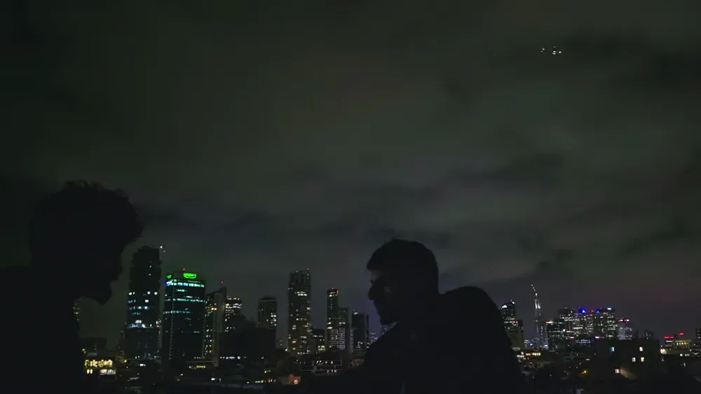 İki kişi, gece şehir ışıklarının önünde siluet hâlinde duruyor.